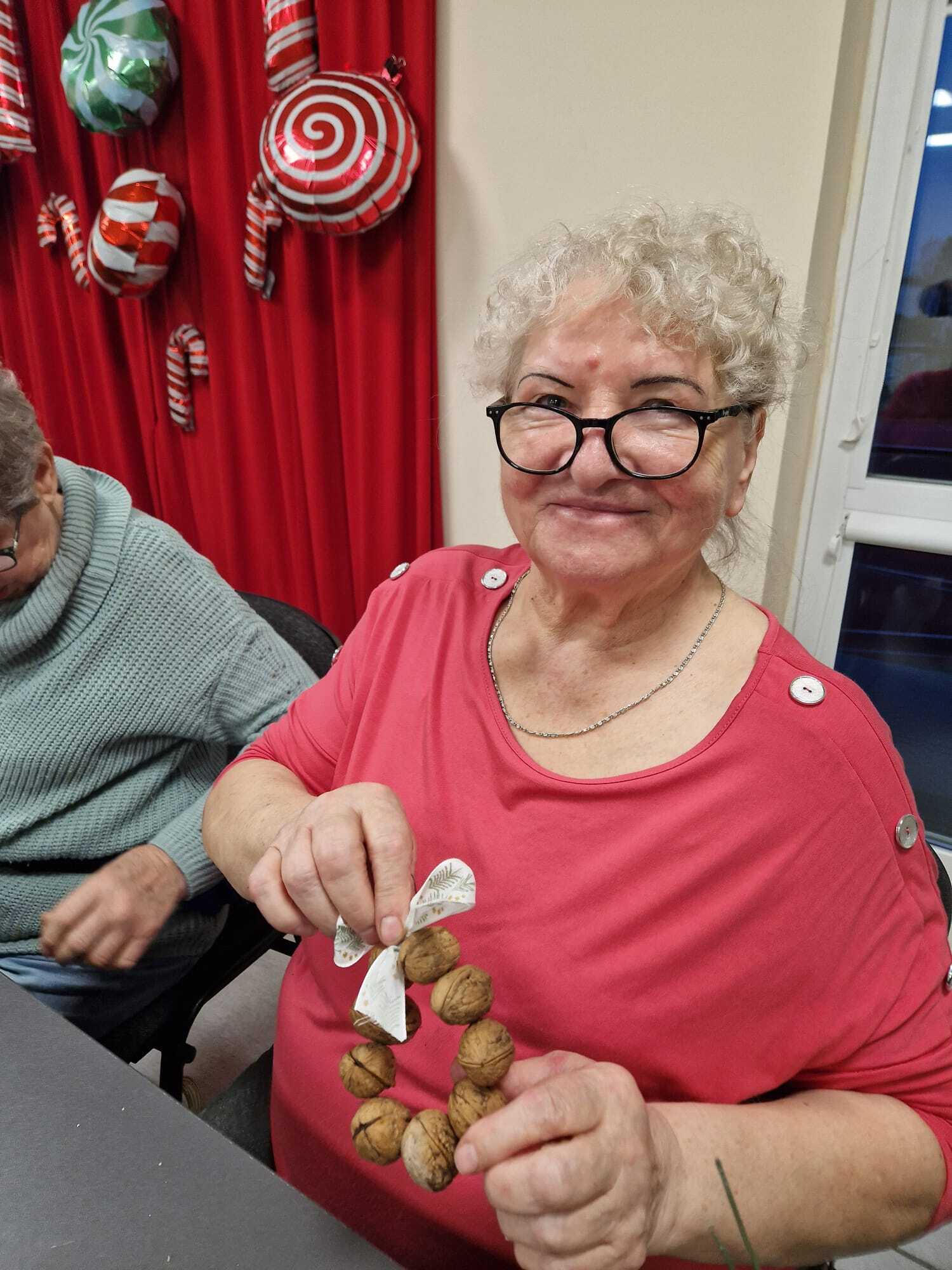 uśmiechnięta uczestniczka warsztatów siedząca przy stole prezentująca w dłoniach gotową ozdobę z orzechów