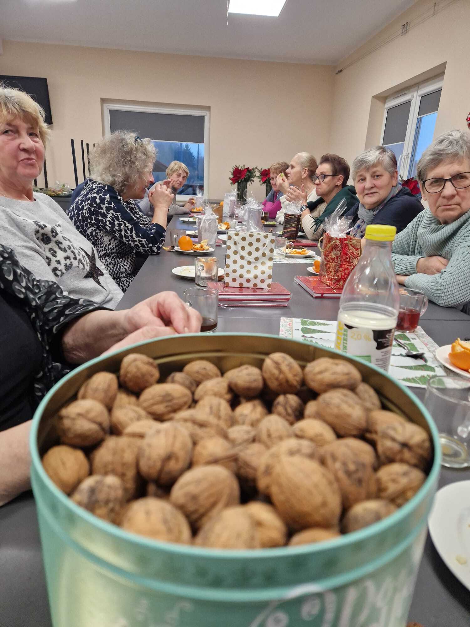 zdjęcie przedstawia metalowe okrągłe pudełko wypełnione całymi orzechami włoskimi, w tle siedzące przy stole uczestniczki warsztatów