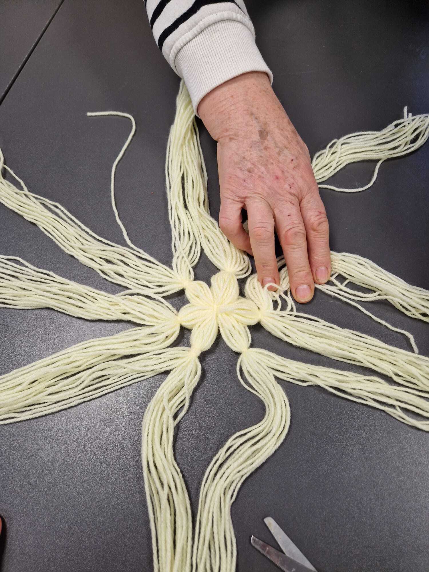 leżąca na blacie stołu ozdoba z włóczki w fazie początkowej tworzenia z dłonią uczestniczki warsztatów